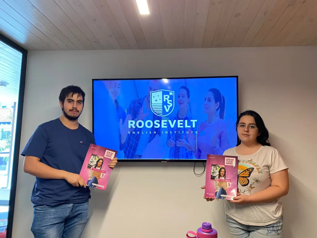 Un joven y una joven posando con sus libros de la serie Speak Your Mind frente a una pantalla con el logo de Roosevelt English Institute.